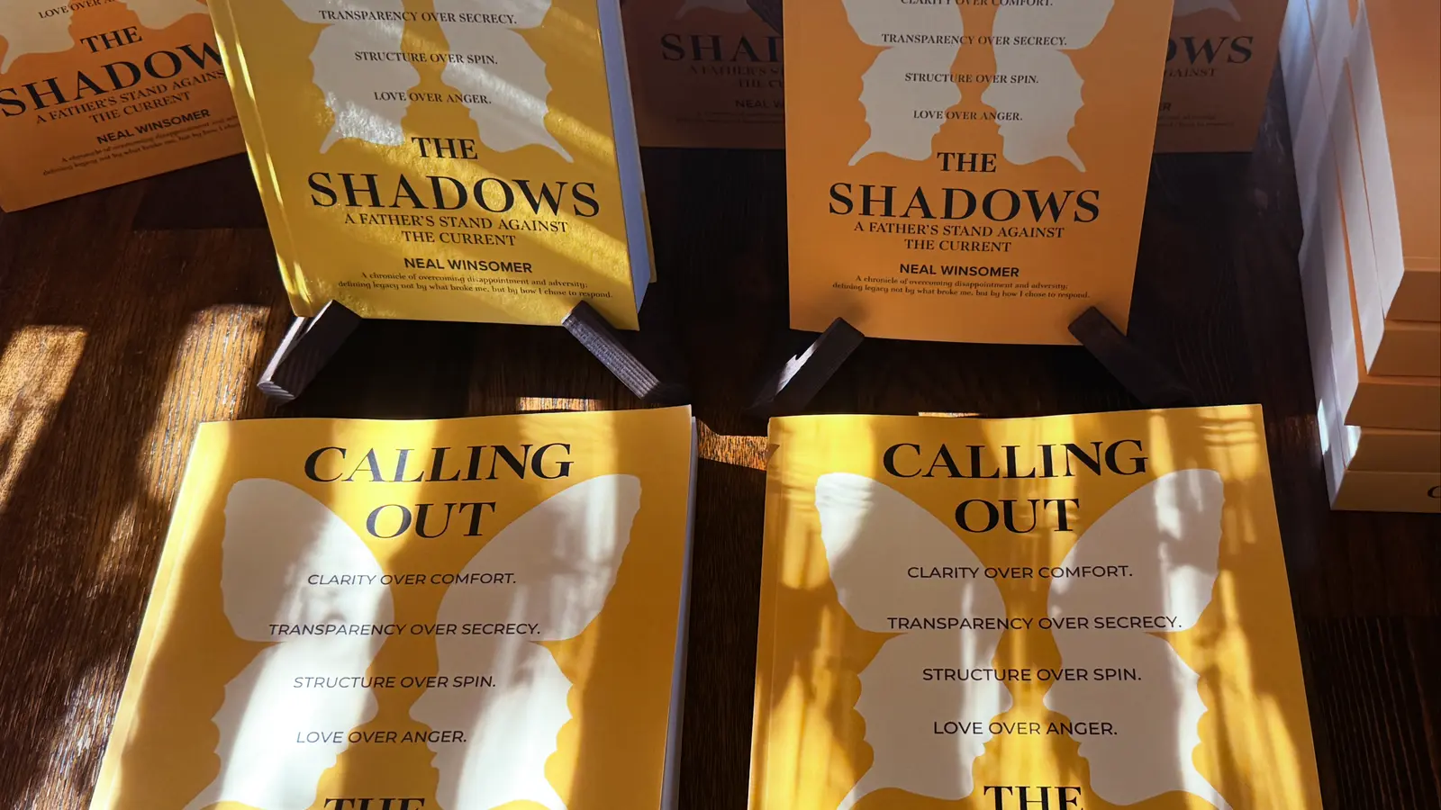 Calling Out the Shadows by Neal Winsomer, hardcover and two-volume large print edition fanned on a wood table in sunlight